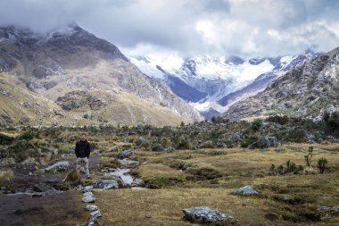 Laguna 69 'a giden yol. Poz veren biri. Etrafında küçük çalılar ve arkasında karlı tepeleri ve bulutları olan büyük dağlar var. Peru, beyaz dağ sırası. Latin Amerika