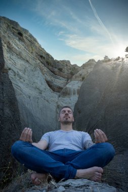 Genç beyaz bir adam mağaraların ortasında meditasyon yapıyor. Yüksek uçurumlar ve güneş ışınları. Dikey fotoğrafçılık
