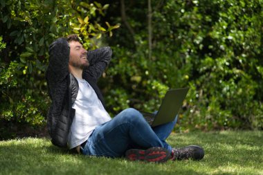 Genç bir adam dizüstü bilgisayarıyla parkta çimlerde otururken elleri başının arkasında sanki esniyor ve rahatlıyor gibi.