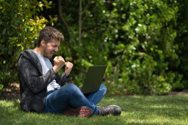 Genç adam parkta oturmuş bilgisayarını izliyor ve elleriyle kutlama yapıyor..