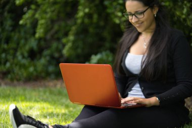 Parkta dizüstü bilgisayarıyla gülümseyen ve ders çalışan esmer bir genç kadın.