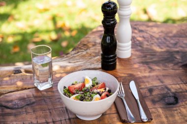 Taze organik salata, yeşil yapraklar, yumurtalar, siyah zeytinler ve ahşap bir kırsal masada, çatal bıçak, bir bardak su, biber ve tuzluk ile servis edilir..