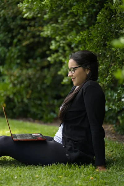 Een vrouw zittend op het gras van een park kijkend naar het scherm van zijn laptop. Natuurlijke omgeving — Stockfoto