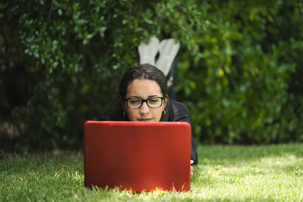 Vrouw liggend op het gras van een park werkend aan haar laptop met haar voeten omhoog. Natuurlijke omgeving — Stockfoto
