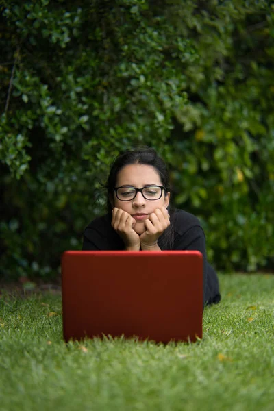 Brunette woman lying down on the grass of a park while watching the screen of her laptop. Vertical Photography — Stock Photo, Image