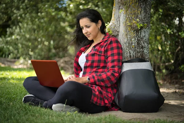 Jonge vrouw zittend onder een boom van een park met haar computer boven haar benen. Natuurlijke omgeving. Technologisch concept — Stockfoto