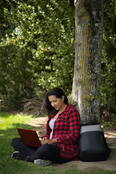 Latijnse vrouw zittend onder een boom van een park met haar computer boven haar benen. Ze heeft haar rugzak naast zich. Technologisch concept. Verticale fotografie — Stockfoto