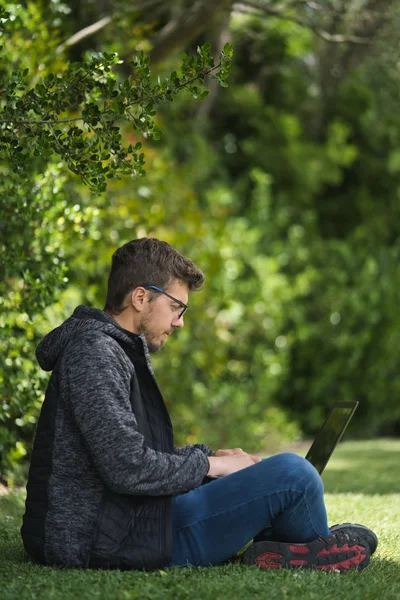 Een blonde man zittend op het gras van een park met gekruiste benen zijn laptop vasthoudend. — Stockfoto