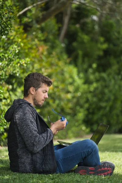 Ένας ξανθός άντρας κάθεται σε ένα πάρκο καθαρίζοντας τα γυαλιά του ενώ κρατάει το λάπτοπ του ανάμεσα στα πόδια του. — Φωτογραφία Αρχείου