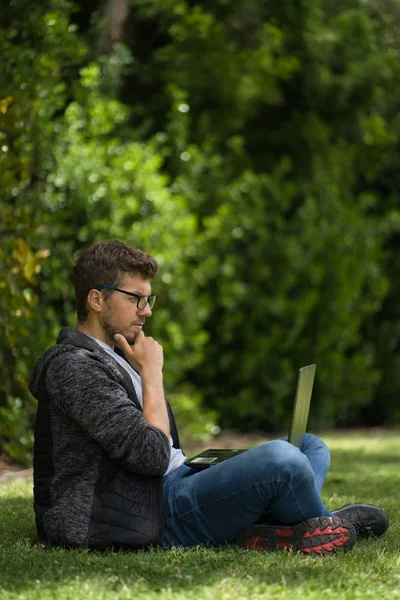 A young blond man is sitting on the grass of a park with his laptop. Natural environment. Vertical photography