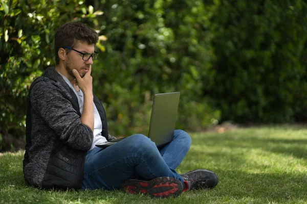 Een jonge man met een bril zit op het gras van een park met zijn laptop. Natuurlijke omgeving. — Stockfoto