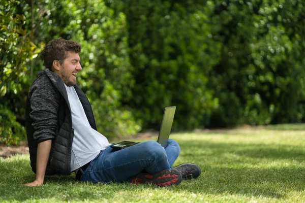 Blanke man lacht terwijl hij naar zijn laptop kijkt. Natuurlijke omgeving — Stockfoto