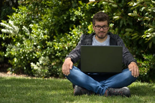 Blanke man zittend op het gras van een park en kijkend naar het scherm van zijn laptop. Natuurlijke omgeving — Stockfoto
