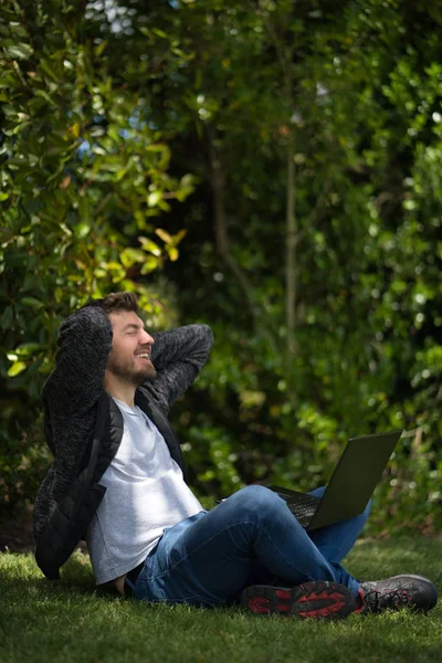 Jongeman zittend op het gras van een park met zijn laptop terwijl hij zijn handen achter zijn hoofd houdt alsof hij zich uitstrekt en ontspant. Natuurlijke omgeving. Verticale fotografie — Stockfoto