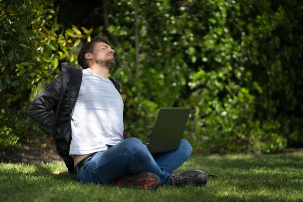 Een jonge blanke man die met zijn computer op het gras van een park zit. Hij klaagt over zijn rugpijn.. — Stockfoto