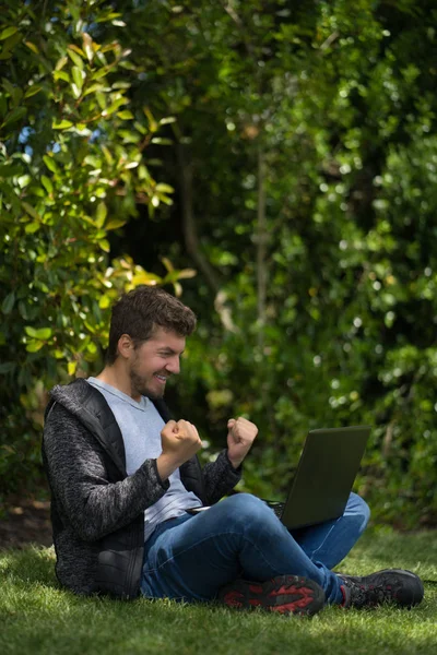 Young man sitting on a park while watching his computer and celebrating furiously with his hands. Natural Environment. — Stock Photo, Image