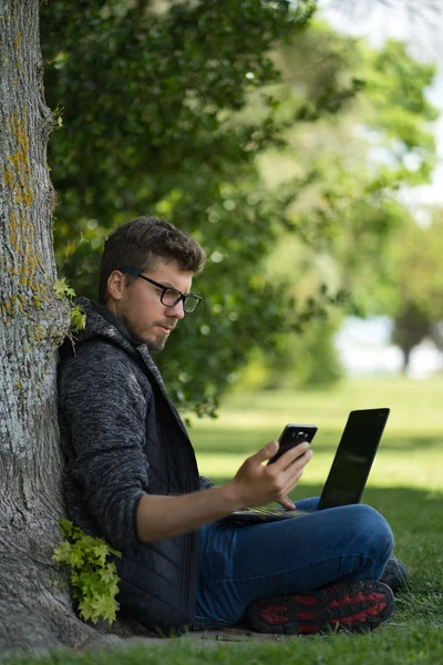 Jonge Kaukasische man werkt aan zijn computer zittend onder een boom terwijl hij tegelijkertijd naar zijn mobiel kijkt. Technologisch concept — Stockfoto