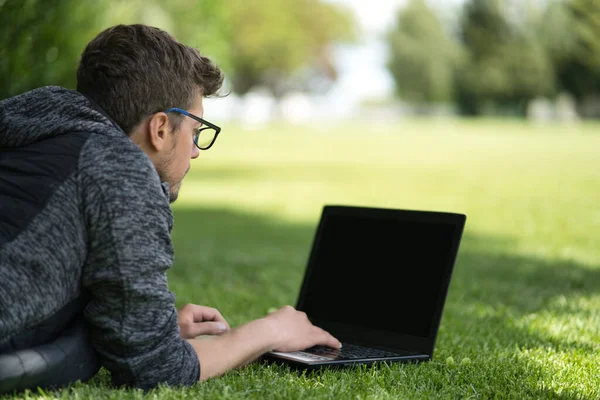 Man liggend op het gras typend op zijn laptop in een prachtig park. Technologisch concept. — Stockfoto