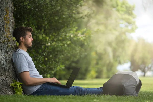 Young man sitting under a tree with his legs stretched and his computer on them — Stock Photo, Image