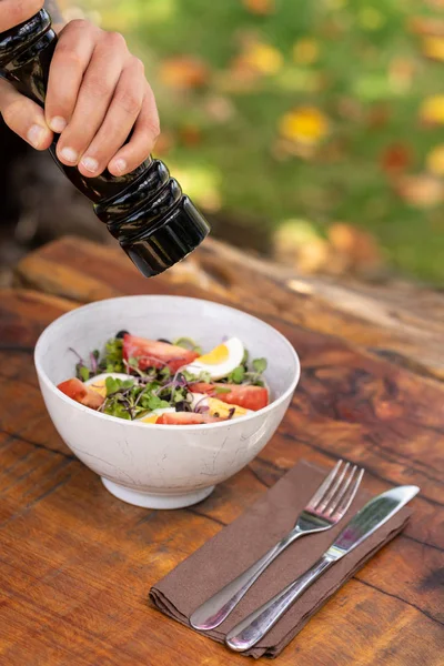 Sazonar a mano con un pimentero negro una ensaladera fresca sobre una mesa de madera con cubiertos. Naturaleza fondo borroso. Fotografía vertical — Foto de Stock