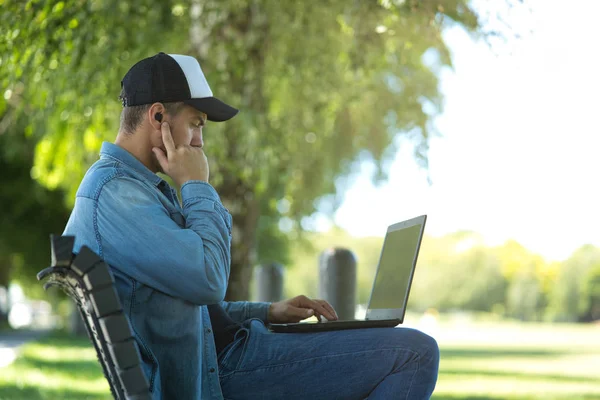 Jongeman met behulp van draadloze oordopjes met zijn laptop op outdoor achtergrond — Stockfoto