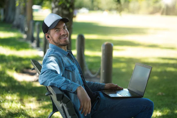 Jongeman met een computer met draadloze oordopjes op een bankje in het park — Stockfoto