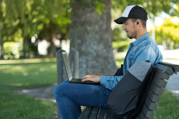 Blanke man luistert naar muziek met draadloze oordopjes en zijn laptop op een bankje in het park — Stockfoto