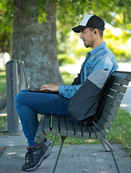 Homme utilisant un ordinateur avec écouteurs sans fil sur un banc de parc — Photo