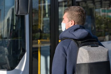 Yüz maskeli ve sırt çantalı bir adam otobüs durağında otobüse binmek üzere. Covid-19 konsepti