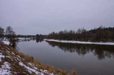 Winter landscape with river and reflections and snow on the Bank on a cloudy day clipart