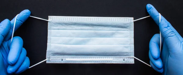 Tek kullanımlık tıbbi maske ellerinde mavi lastik eldivenlerle siyah arka planda yer alan. Kavram: Coronavirüsü durdurun