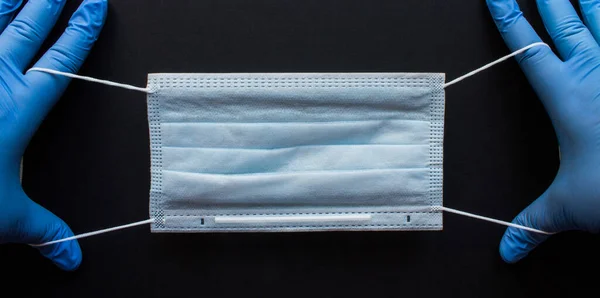 Tek kullanımlık tıbbi maske ellerinde mavi lastik eldivenlerle siyah arka planda yer alan. Kavram: Coronavirüsü durdurun