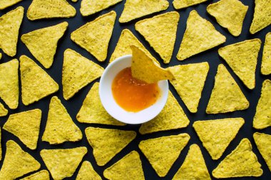 Siyah bir zemine yayılmış mısır cipsleri ve ortasında sos olan beyaz bir kupa.