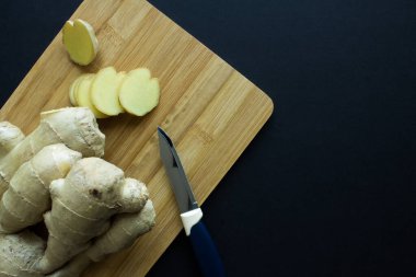 Ahşap mutfak tahtasının üzerinde dilimlenmiş parçalarla taze zencefil kökü ve siyah bir arka plana yakın bir bıçak.