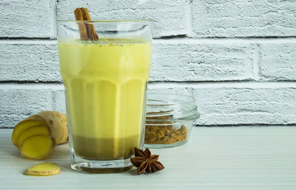 Golden milk in a clear glass and ginger root with a cinnamon stick against a white brick wall. The trend is healthy eating.