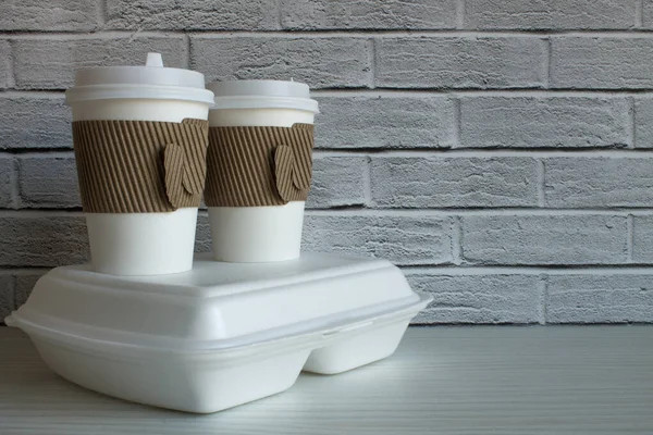 Two disposable coffee cups and a white container with food in close-up against a brick wall. The concept of food delivery takeaway