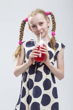 Komik ve meraklı gülümseyen Pigtails puantiyeli elbise kırmızı suyu fincan ile ayakta beyaz kızla portresi.