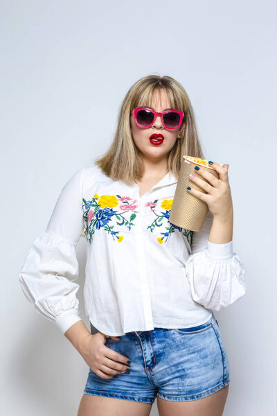 Portrait of Sensual Caucasian Female Blond Girl In Shorts and Red Sunglasses Holding Popcorn Bucket and Posing Against White. Vertical Image Composition