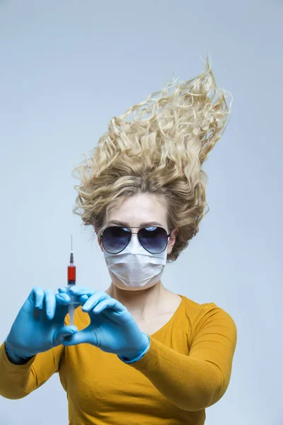 Portrait of Caucasian Woman with Lifted Hair. Wearing Flu Virus Mask For Viral Prevention With Black Glasses.Posing in Protective Gloves with Syringe. Against Gray. Vertical Image Orientation