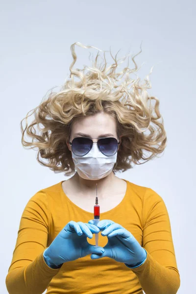 Portrait of Caucasian Woman with Lifted Hair. Wearing Flu Virus Mask For Viral Prevention With Black Glasses.Posing in Protective Gloves with Syringe. Against Gray. Vertical Image