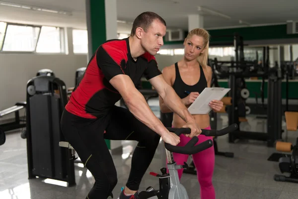 Uomo treno bicicletta sulla macchina con personal trainer — Foto Stock