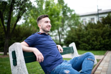 Park 'ın dışında bankta oturan, kendinden emin ve başarılı mavi gömlekli genç bir adamın portresi.