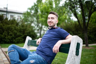 Park 'ın dışında bankta oturan, kendinden emin ve başarılı mavi gömlekli genç bir adamın portresi.