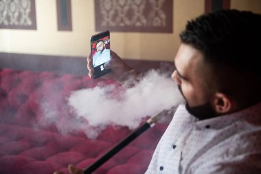 Arka planda renkli duvarları olan ve telefonla selfie çeken bir adam kafede Türk nargilesi içiyor.