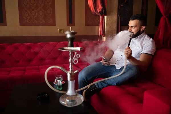 在阿拉伯餐馆抽烟的年轻人 男人从胡卡呼出烟雾并通过电话发短信 — 图库照片