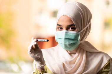 Cerrahi Maskeli ve Eldivenli Müslüman Kadın Doktor elinde Covid-19 olarak imzalanan bir kan testi tüpü tutuyor.