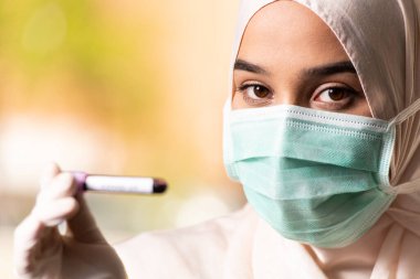 Cerrahi Maskeli ve Eldivenli Müslüman Kadın Doktor elinde Covid-19 olarak imzalanan bir kan testi tüpü tutuyor.