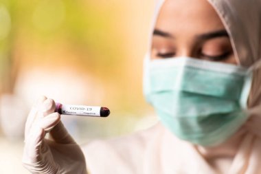 Maskeli ve Eldivenli Müslüman Kadın Doktor elinde Covid-19 olarak imzalanmış bir kan testi tüpü tutuyor.