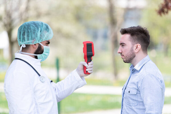 Doctor Man Use Infrared Forehead Thermometer Gun to Check Body Temperature for Virus Covid-19 Symptoms With the Isolation Gown or Protective Suits and Surgical Face Masks Outdoors in Park