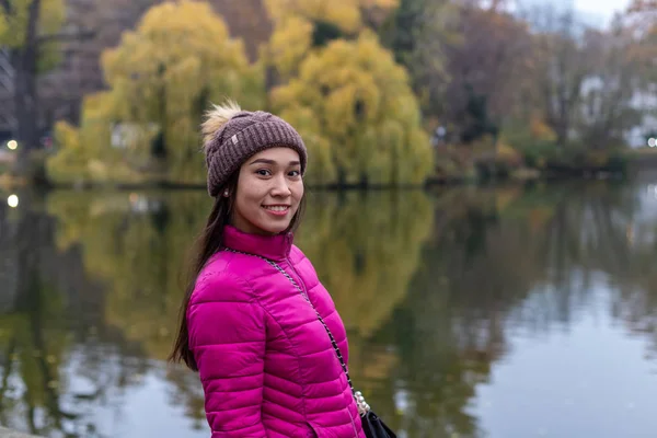 Smiling asian girl with freckles and unusual look. Attractive girl in a pink jacket and Knitted hat. On a blurred background pond and trees